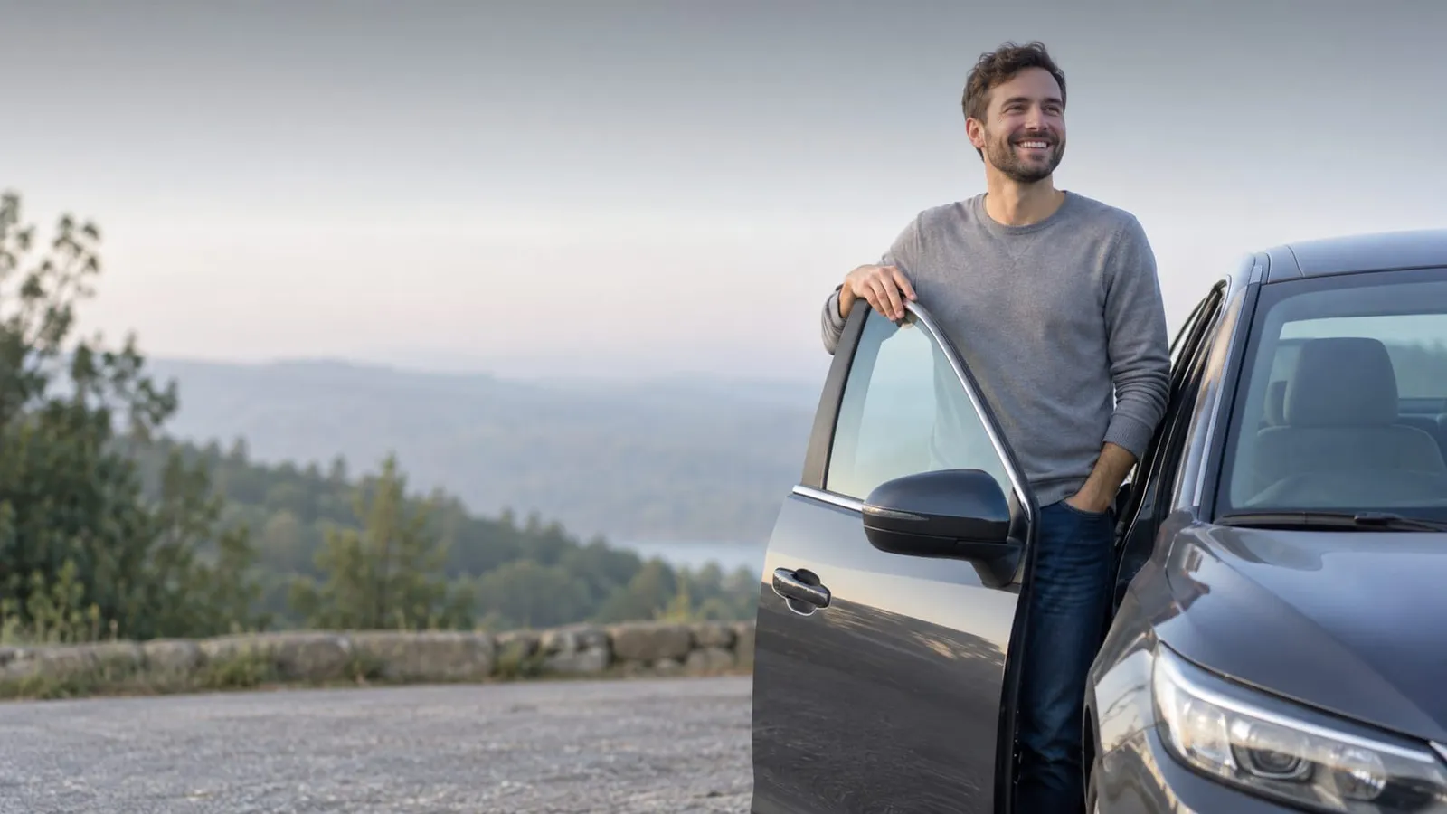 A smiling man leans on the open door of a dark car at an overlook in soft morning light