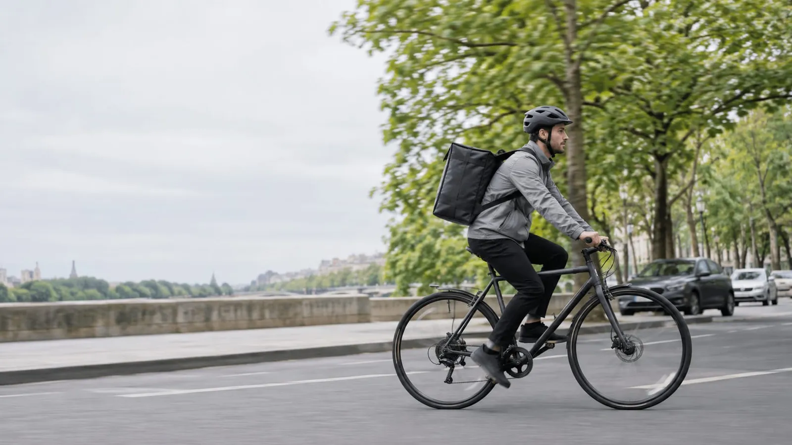 A cyclist with a black insulated delivery backpack rides along a tree-lined riverside road
