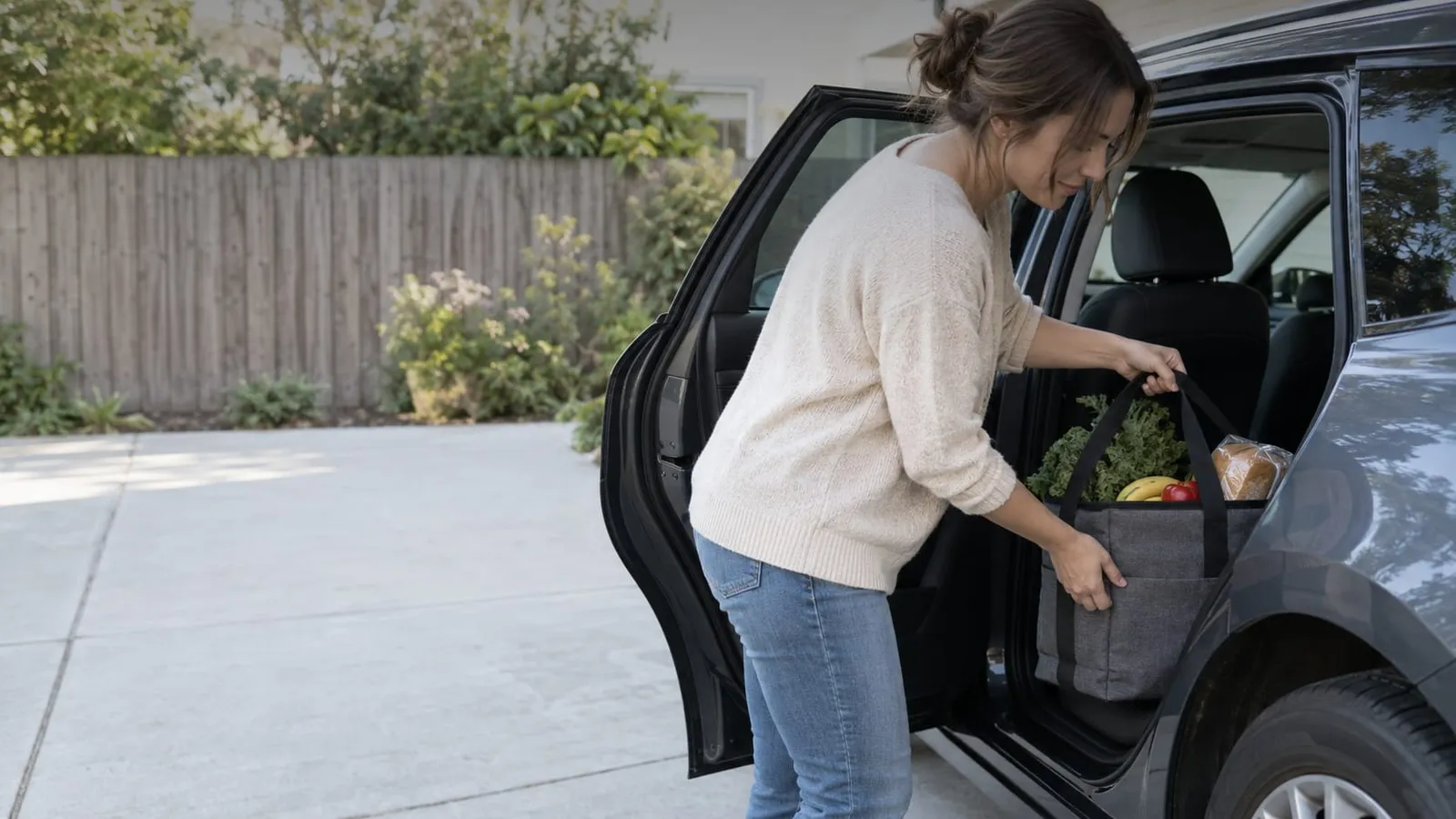 A woman leans into the back seat of a car to lift an insulated grocery bag with greens, bananas and bread