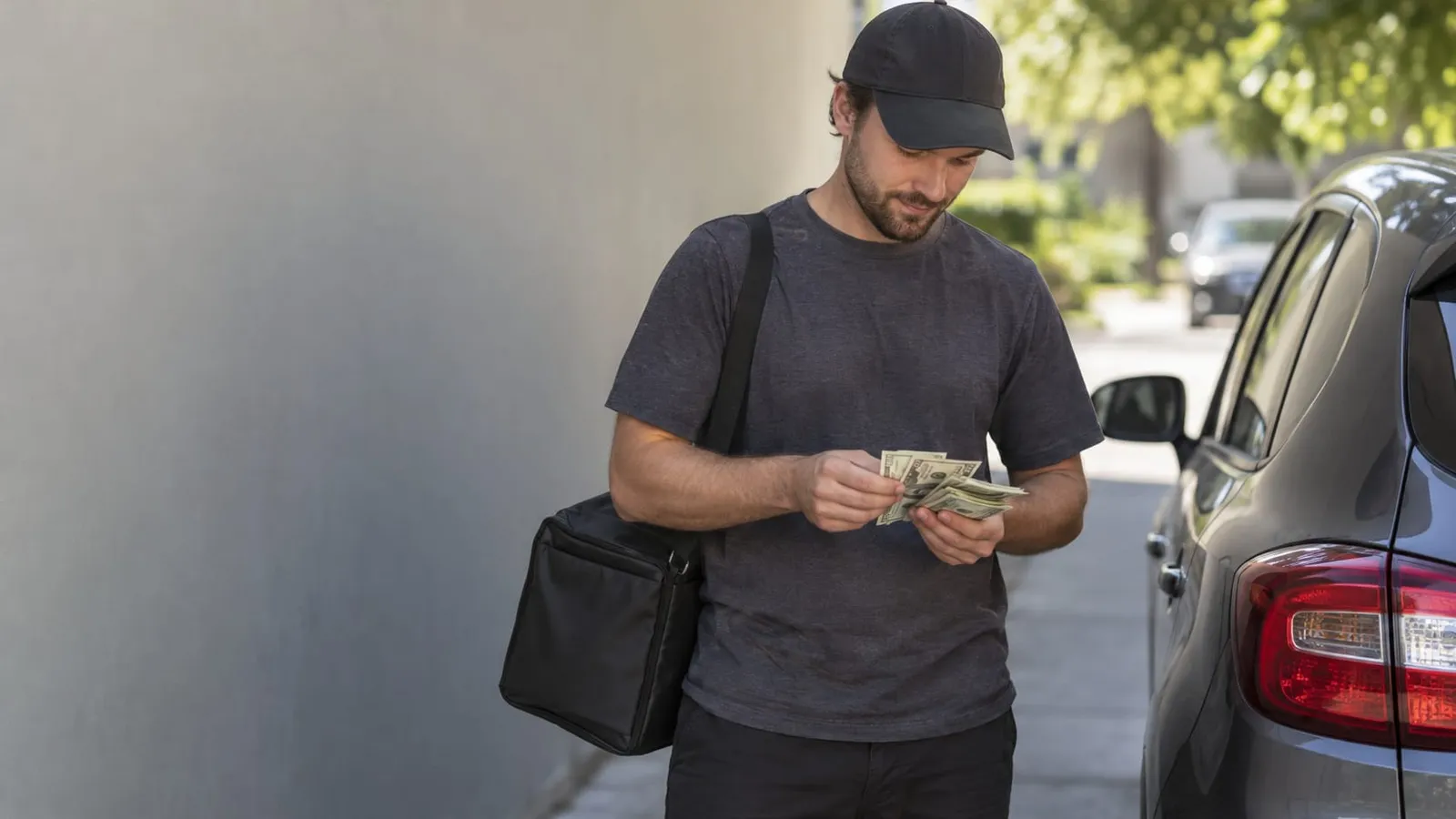 A man in a black cap with a delivery bag on his shoulder counts cash next to a gray car
