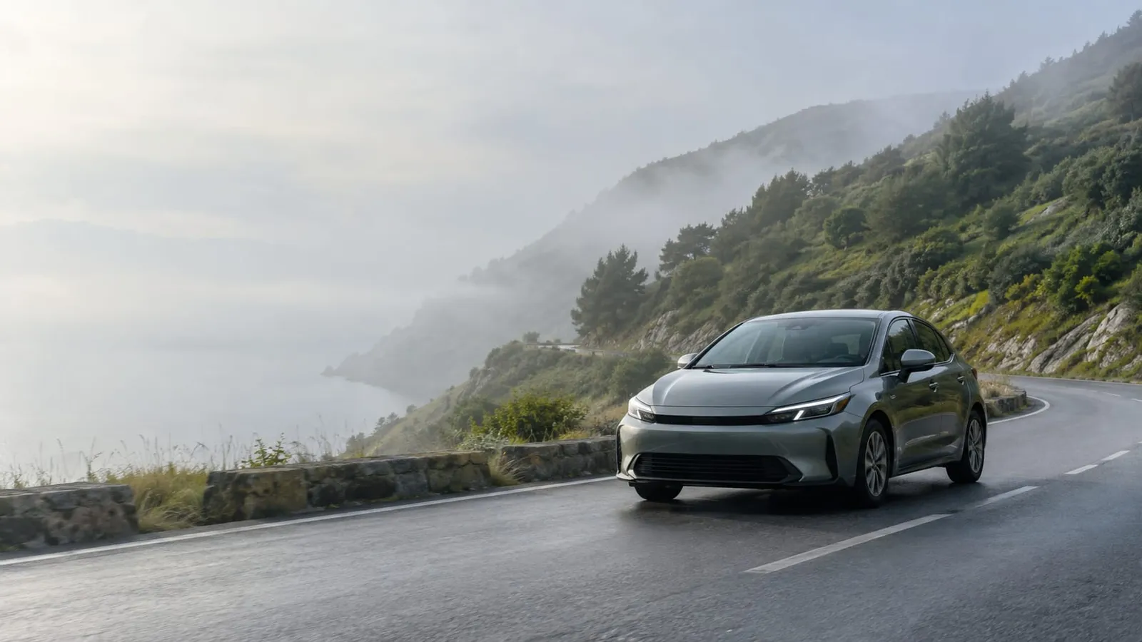 A gray sedan driving along a misty coastal hillside road in the morning