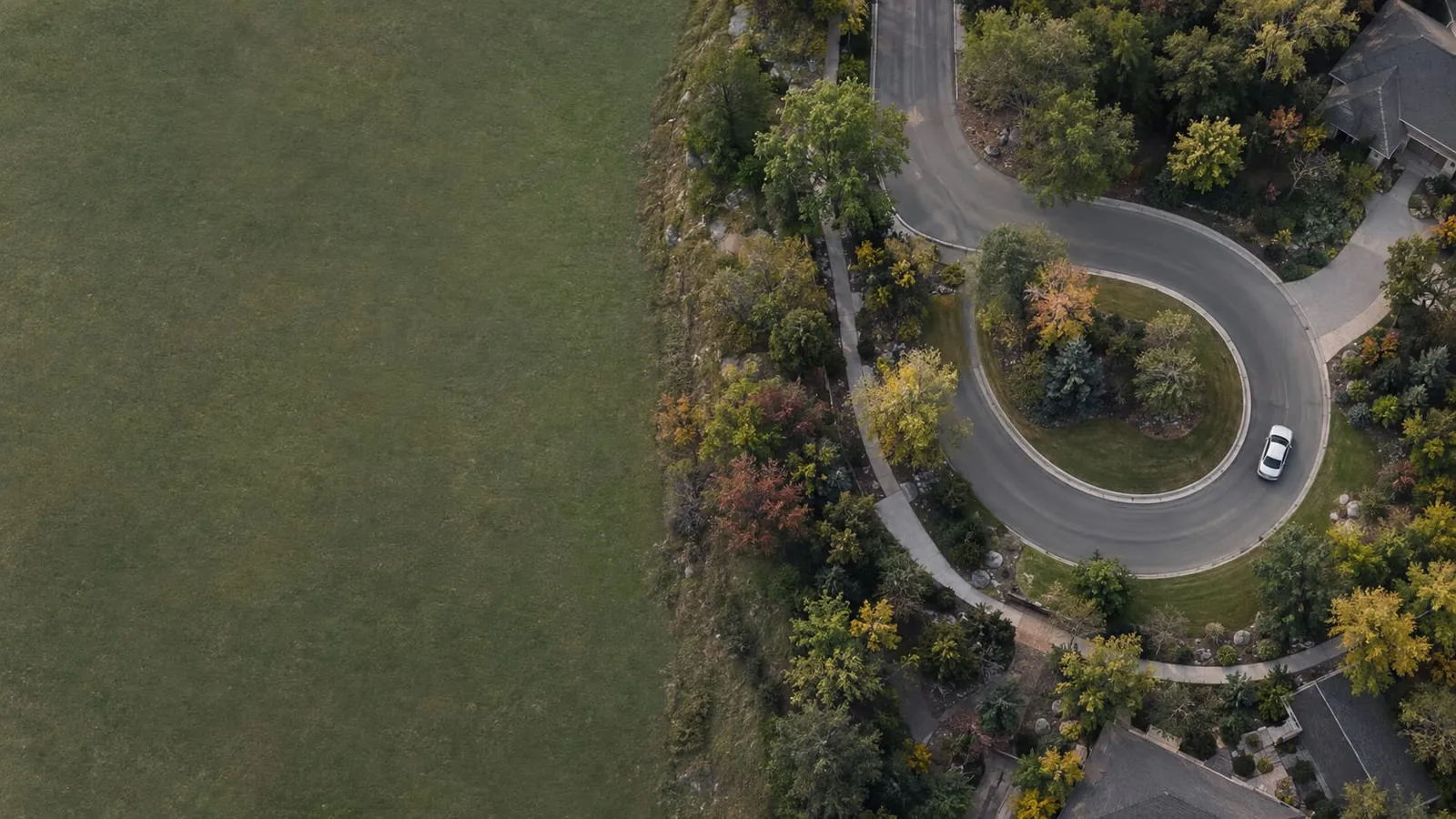 Aerial view of a white car driving around a looping road through trees and houses beside a large green field