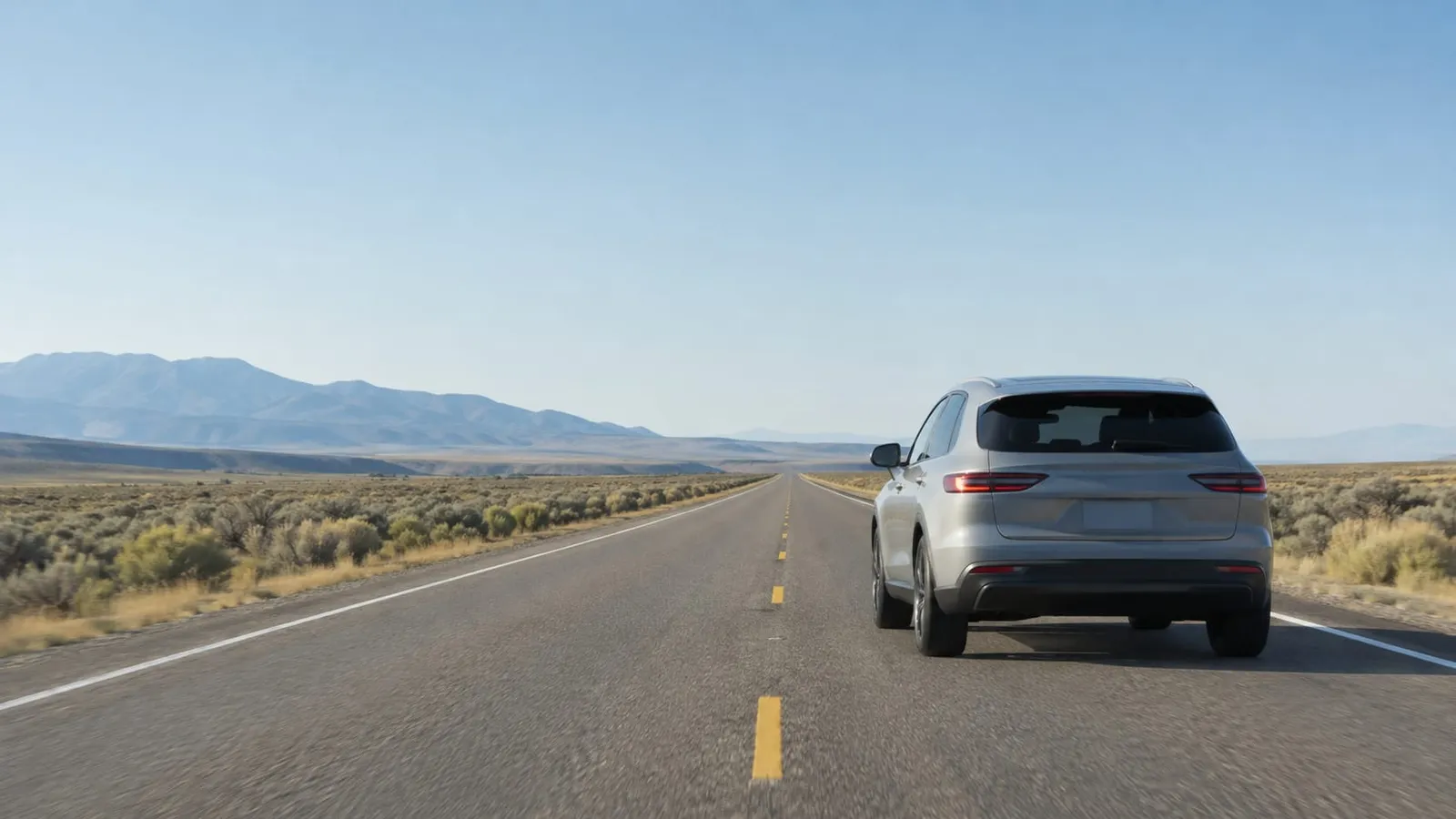A silver SUV seen from behind on an empty two-lane highway with mountains in the distance