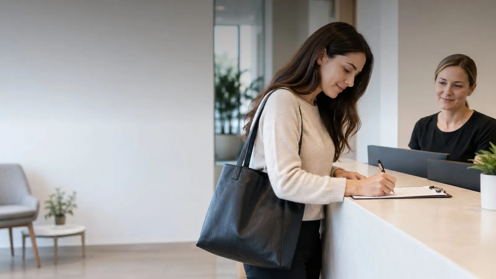 A woman with a black tote bag fills in a form on a clipboard at a reception desk while a receptionist smiles