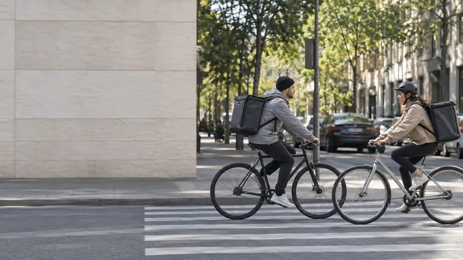 Two couriers with insulated delivery backpacks riding bicycles toward each other across a crosswalk on a tree-lined city street