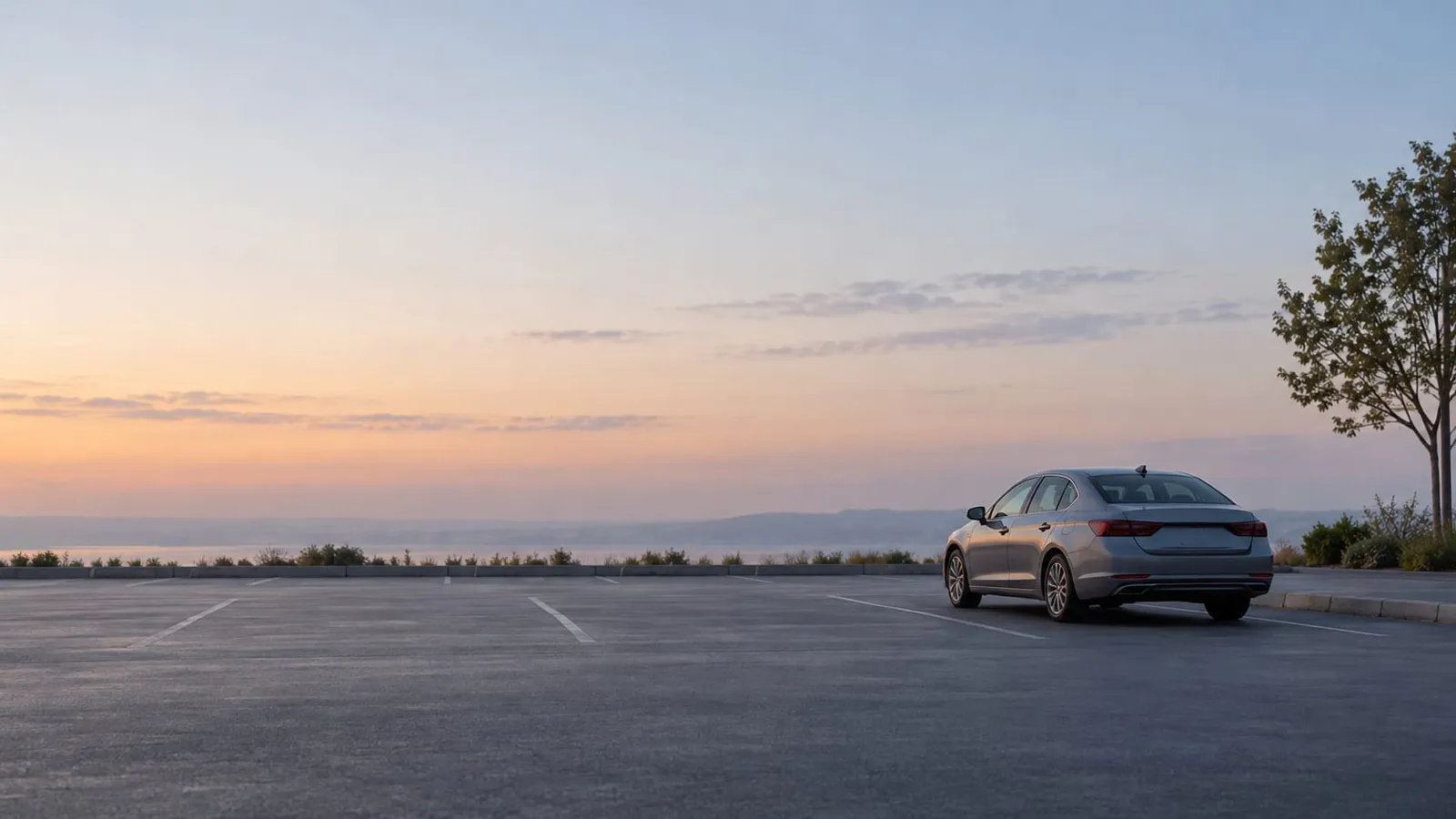 A lone gray sedan parked in an empty parking lot at sunrise with a hazy sky and water in the distance