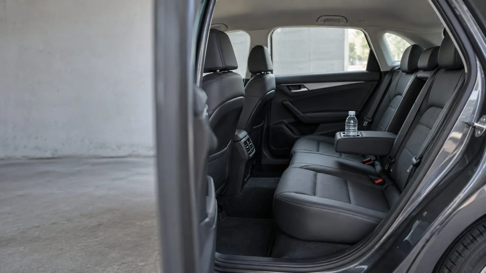 The leather back seat of a gray car seen through an open rear door, with an armrest and a bottle of water in the cup holder