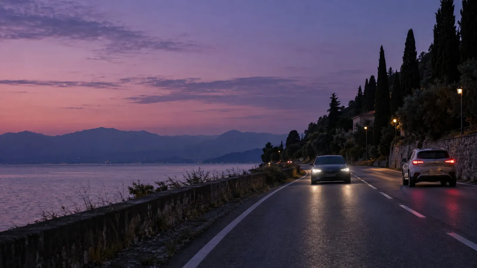 Two cars driving on a coastal road at dusk with a purple sky, mountains and a calm sea in the background